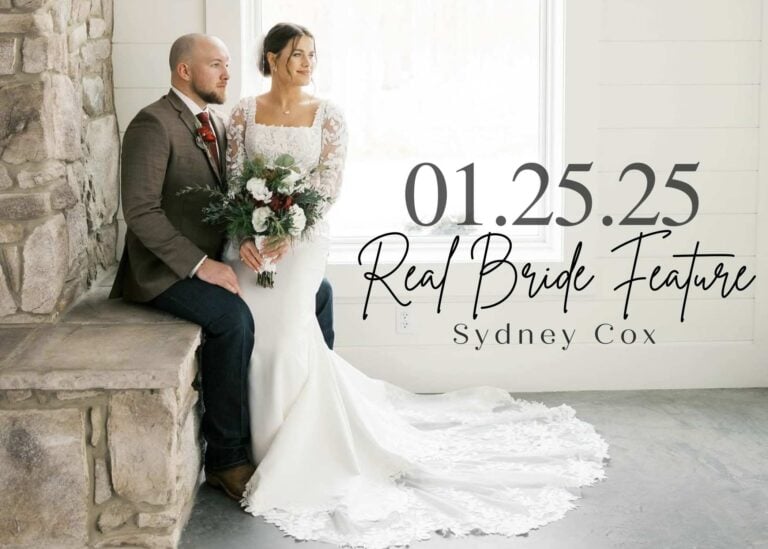 Bride Sydney Cox sitting beside her groom on a stone ledge, wearing a fitted long-sleeve lace wedding gown with a dramatic scalloped train and holding a winter-inspired bouquet, framed by a bright window.
