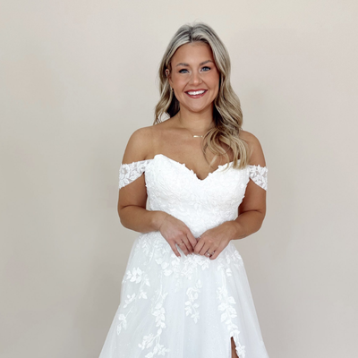 A woman in a white off-shoulder wedding dress with floral designs, smiling at the camera, showcasing a stunning option for any wedding gown budget.