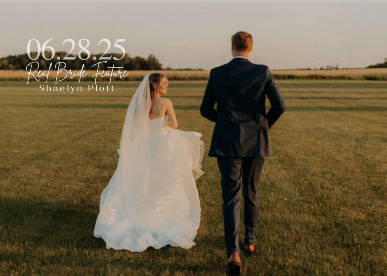 Bride Shaelyn Plott walking across a sunlit field in a flowing wedding gown and veil, holding her train while looking back at the camera, with her groom walking beside her.