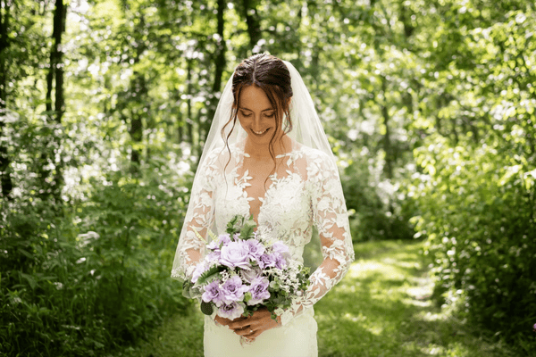 Bridal portrait of a woman in a lace wedding dress showcasing bridal dress customizations, holding a purple flower bouquet in a green forest.