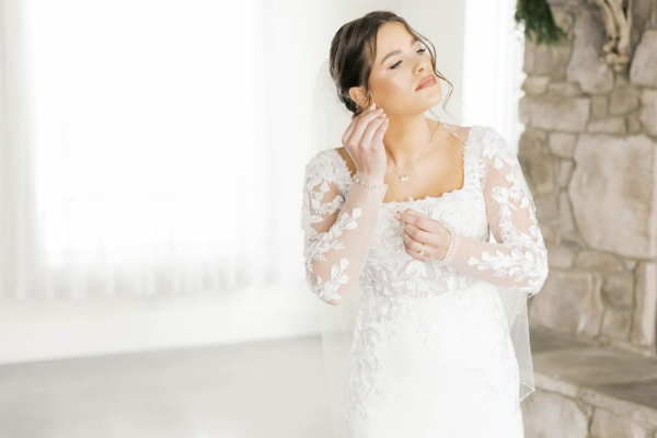 A bride in a white lace wedding dress thoughtfully adjusts her earrings, showcasing her wedding dress accessories.