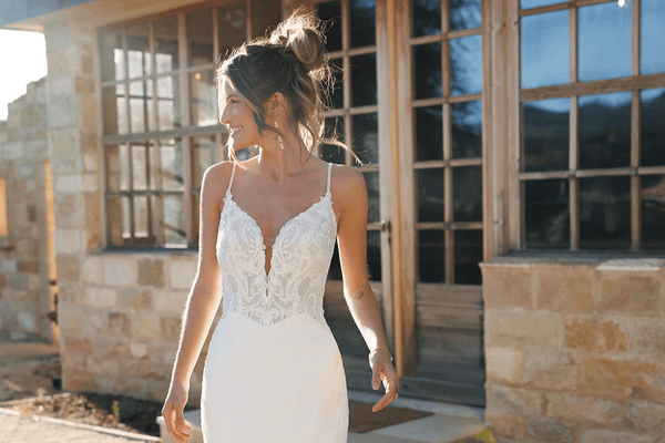 A woman in a beautiful white wedding dress with lace details, smiling outside a stone building.