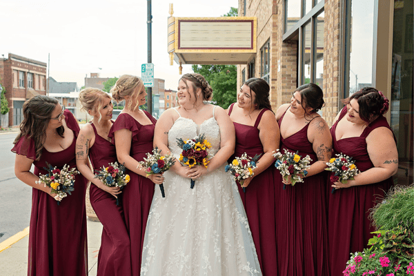 A bride in a white dress and bridesmaids in burgundy dresses, smiling and holding flower bouquets, showcasing perfect bridesmaid dress coordination.