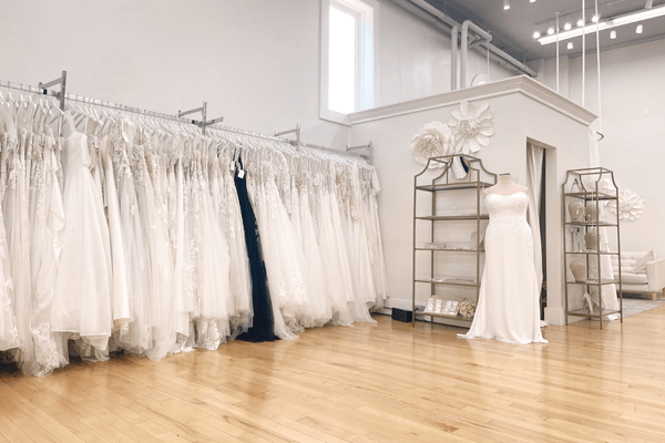 A bridal shop with rows of white dresses hanging, a display mannequin, and options for a custom wedding dress. The floor is wooden.