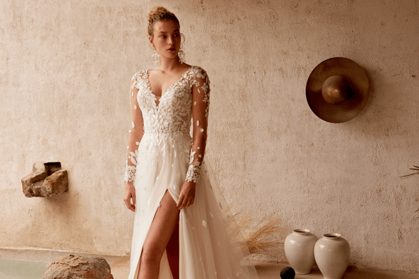 A woman in a white wedding dress with floral details, standing near a stone wall and pottery.