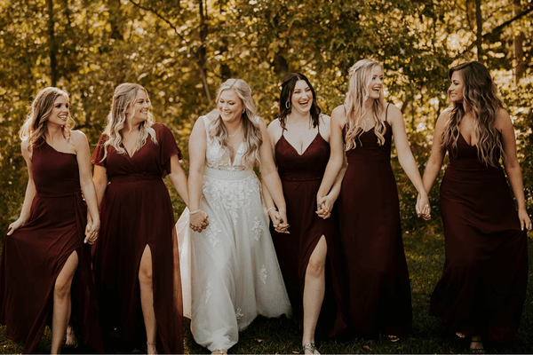 Six bridesmaids in burgundy dresses and the bride in a white wedding gown, holding hands in a green park.