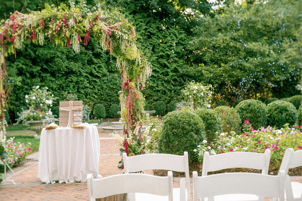A decorated arch covered with greenery and flowers stands in a garden, with white chairs set up for a wedding.