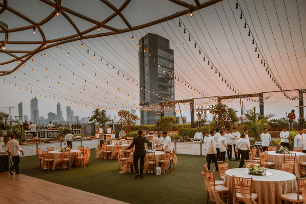 An outdoor wedding setup with round tables, chairs, and servers under a tent at sunset.