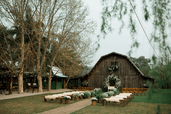 A rustic outdoor wedding setup with wooden benches, greenery, and a charming barn in the background.