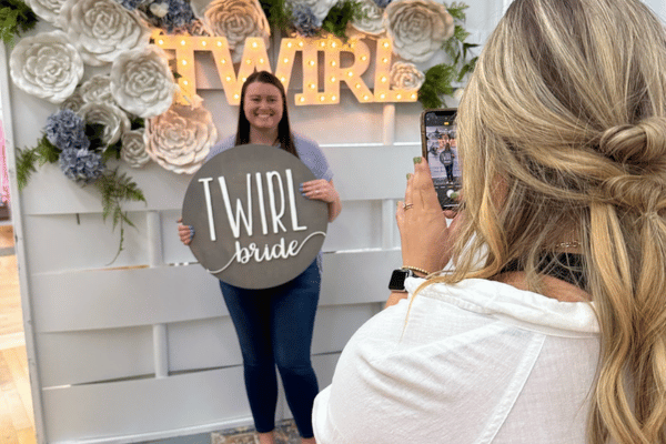 Bride in a shirt and jeans holding a sign that says “Twirl Bride,” while a lady in white takes her photo