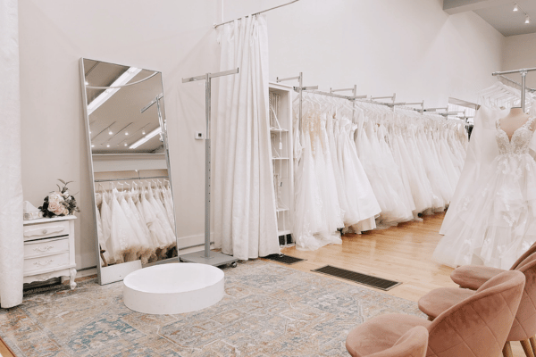 Bridal fitting area with a mirror and platform, seating in front, a curtain to the side, and racks of wedding dresses nearby