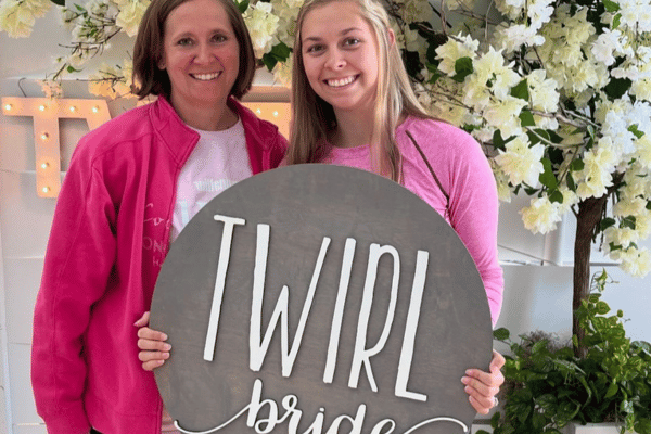 Bride and her mom smiling, bride holding a “Twirl Bride” sign, with flowers in the background