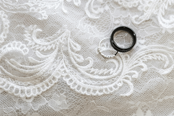 Silver and black wedding bands rest on intricate white lace, showcasing delicate floral patterns. The scene conveys elegance and romance.