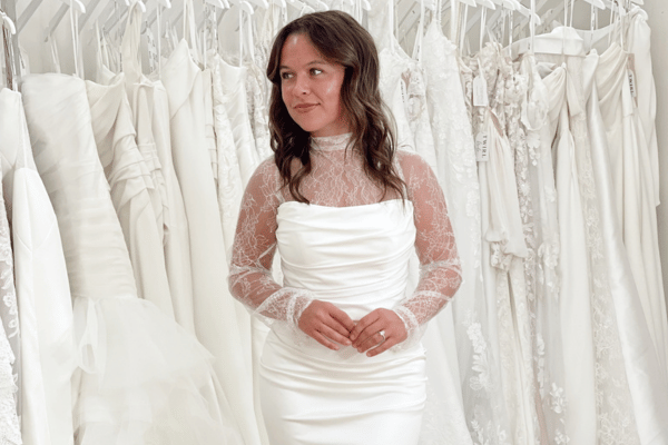 A bride stands among white wedding dresses, wearing a satin gown with lace sleeves. The scene conveys elegance and anticipation in a bridal shop.
