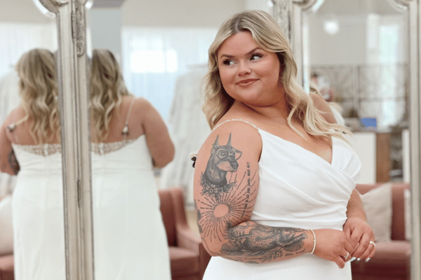 A bride smiles confidently, showcasing her wedding dress. She stands in front of a mirror, reflecting her from multiple angles. The setting appears elegant and bright, with soft lighting and neutral tones, conveying warmth and self-assurance.