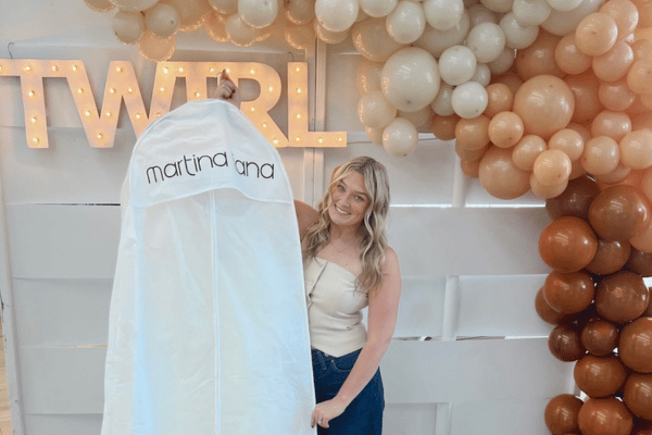 A smiling bride holds a garment bag labeled "martina lana" in front of illuminated "TWIRL" letters. Beige balloons form an arch, creating a festive atmosphere.