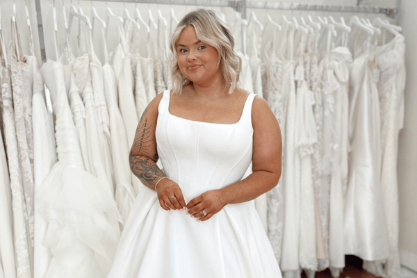 A bride in a white wedding gown stands confidently in a bridal shop, surrounded by rows of hanging dresses. She smiles warmly, exuding joy and excitement.