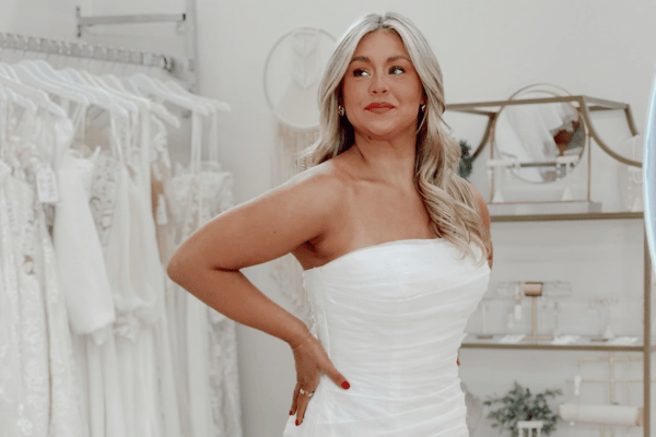 A bride smiles confidently while trying on a strapless white wedding dress in a bridal shop. Racks of dresses are in the background, creating an elegant and joyful atmosphere.