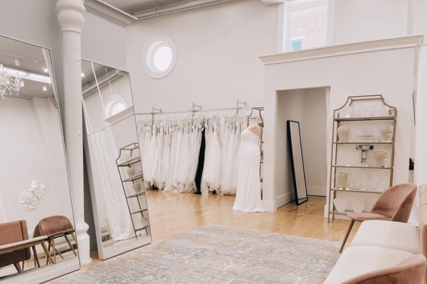 Bright bridal boutique with elegant gowns on racks, a mannequin in a white dress, large mirrors, soft seating, and warm wooden floors.
