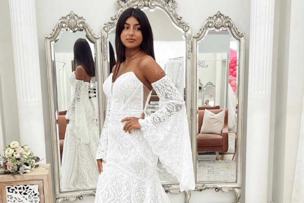A bride wearing a white, lace wedding dress with off-the-shoulder sleeves stands in front of an ornate mirror. The elegant room has soft pastel tones.