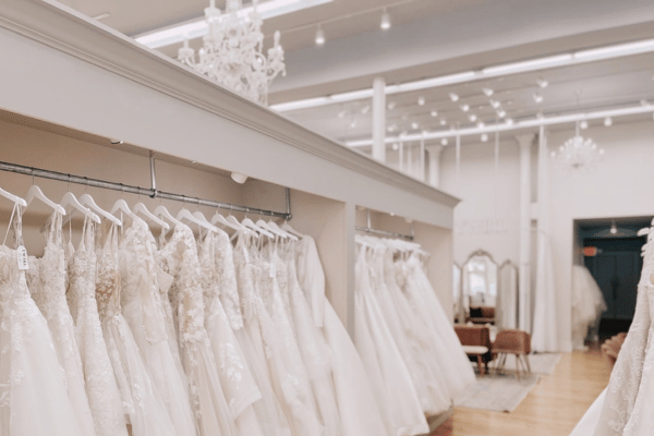 A bridal boutique showcasing a row of elegant white wedding dresses on hangers, set in a spacious, well-lit room with chandeliers and a cozy fitting area.