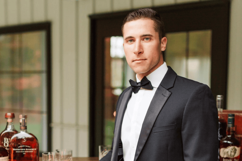 A man in a black tuxedo with a bow tie stands confidently beside a table with bottles of liquor. The setting is sophisticated and formal.