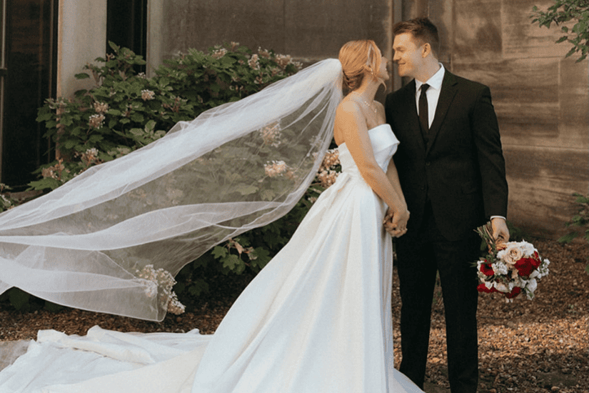 Bride and groom stand smiling, holding hands. The bride's veil flows elegantly, and the groom holds a bouquet. They are surrounded by greenery.