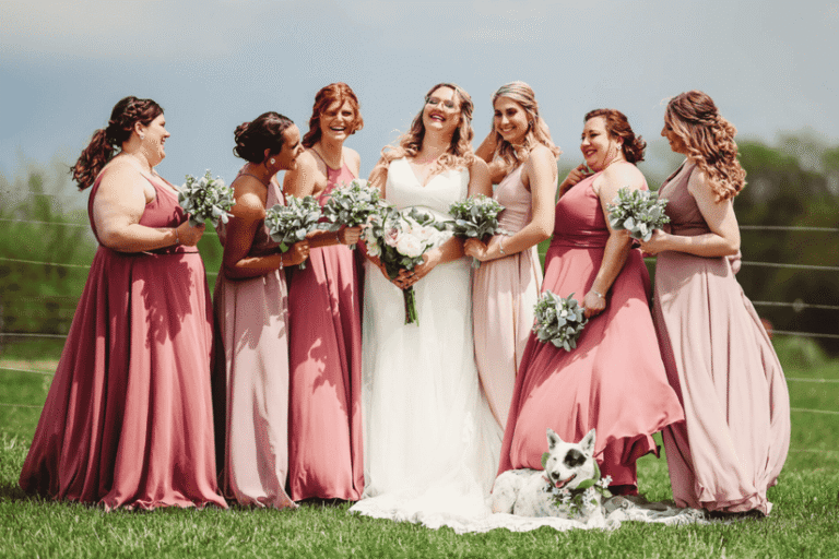 A joyful bride stands with six bridesmaids in pink dresses, holding bouquets. They smile together outdoors, with a happy dog sitting in front on the grass.