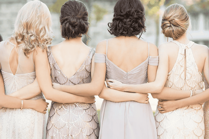 Four women in elegant dresses stand with their backs to the camera, arms around each other. The mood is joyful and supportive, set outdoors.