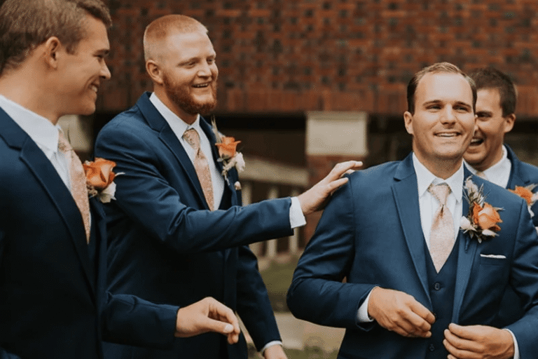 Four groomsmen in matching blue suits and orange boutonnieres share a joyful moment, smiling and laughing together outdoors before a brick wall.
