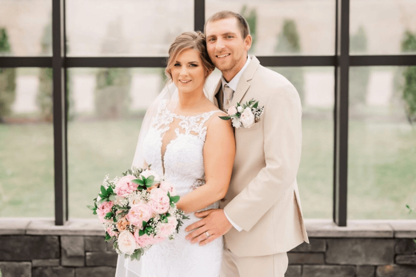A bride in a lace gown with a pink and white bouquet stands beside a groom in a beige suit, both smiling warmly in a bright, elegant setting.