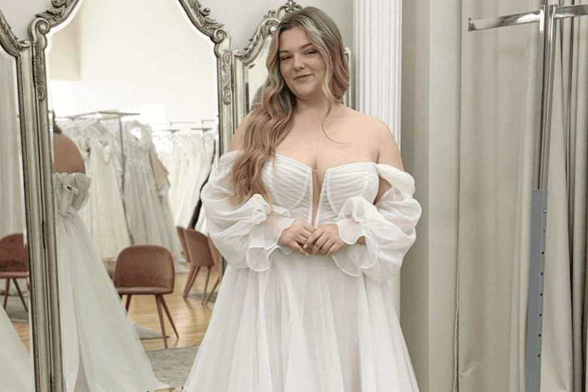 A smiling woman in a bridal boutique wears an off-shoulder, white wedding dress, with mirrors and more dresses visible in the soft-lit background.