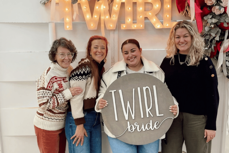 Four women smiling warmly stand together indoors, one holding a round sign reading "TWIRL bride." Behind them, lit letters spell "TWIRL," creating a joyful, celebratory atmosphere.