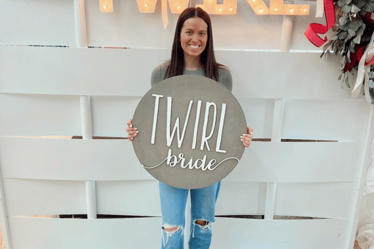 Smiling woman holds a round sign saying "TWIRL bride" in white letters. She's standing in front of a white backdrop with lit letters and greenery.