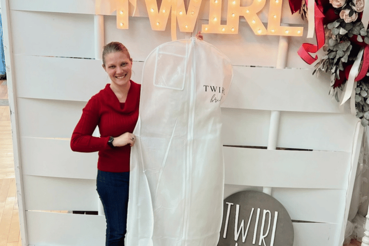 A person in a red sweater and jeans smiles while holding a white garment bag from "Twirl" in front of a lit sign and floral decorations, conveying joy.