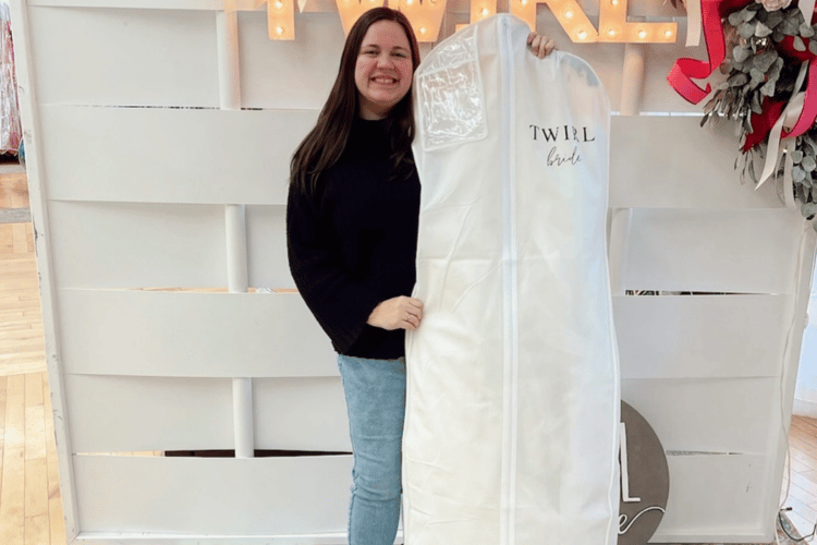 Smiling woman holding a white garment bag labeled "Twirl Bridal" in a bright, warmly lit space with a festive backdrop. Joyful and celebratory mood.