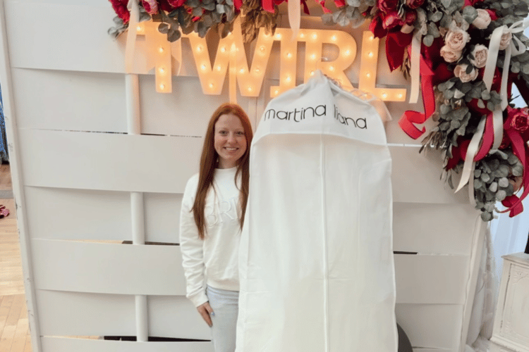 A woman smiles while standing next to a bag labeled "martina liana" in front of a "TWIRL" sign with lights. Floral decorations add a festive touch.