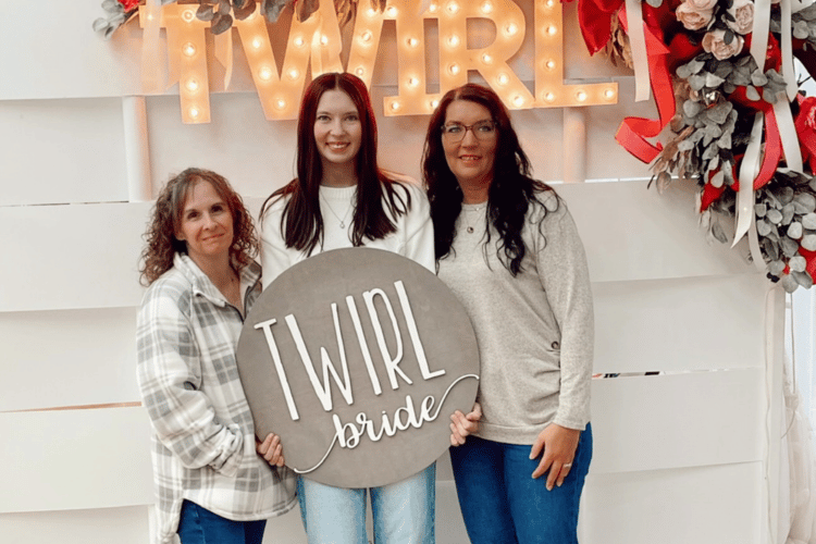 Three women smiling in front of a "TWIRL" sign with lights, holding a circular sign reading "TWIRL bride." They stand before a floral and ribbon display.