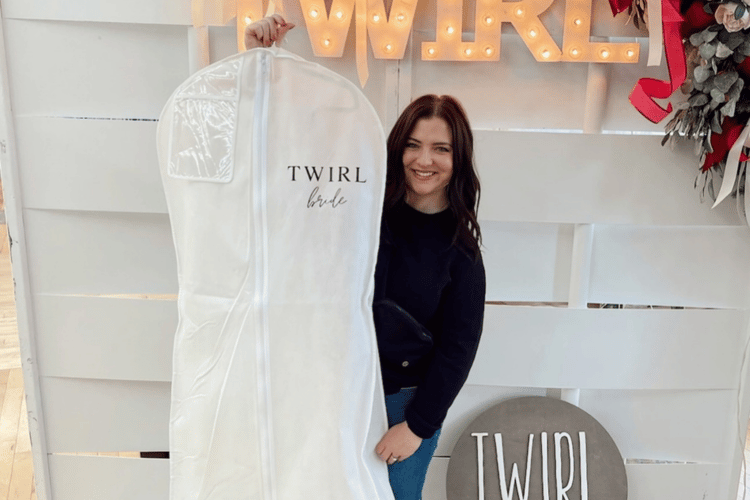 Smiling woman holding a white garment bag labeled "Twirl Bride" in front of a lit sign spelling "TWIRL." Festive decor adds a joyful tone.