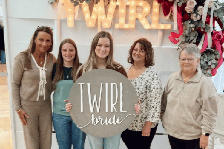 Five women stand smiling in front of a lighted "TWIRL" sign. The central woman holds a circular "TWIRL bride" sign. The atmosphere is joyful and celebratory.