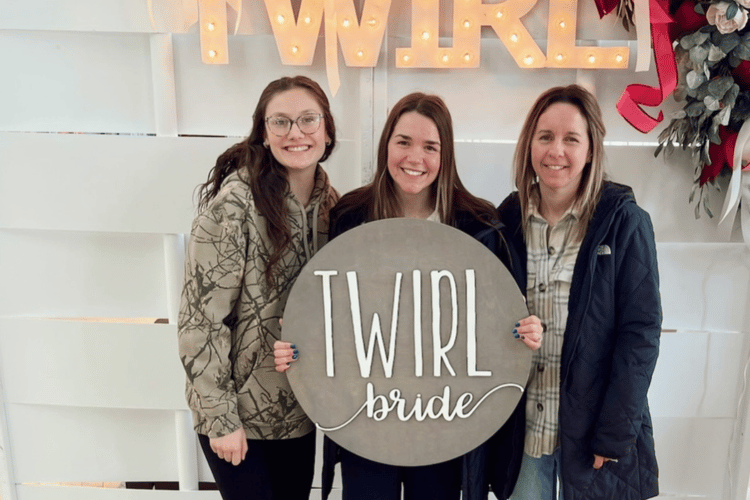 Three women smiling, holding a round sign that reads "TWIRL bride" in front of a lit "TWIRL" sign. Casual, joyful atmosphere.