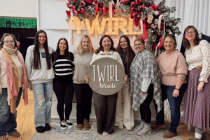 A group of nine women, smiling, stands in front of a floral backdrop with a lit "TWIRL" sign. The woman in the center holds a "TWIRL bride" sign.