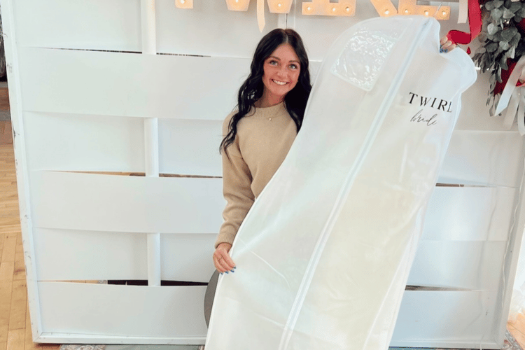 Young woman smiles holding a long white garment bag with "TWIRL bride" text, in a bright space with wood flooring and white decor. Joyful atmosphere.