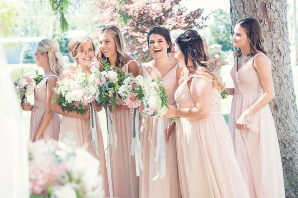 A group of bridesmaids wearing blush pink floor-length dresses stand outdoors holding bouquets of white and pink flowers with greenery.