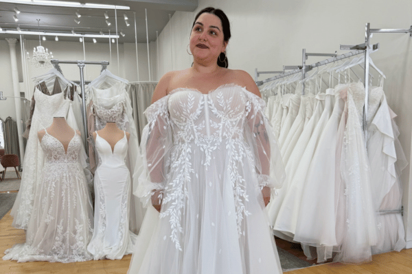 Bride wearing an off-the-shoulder wedding dress with floral lace detailing in a bridal boutique.