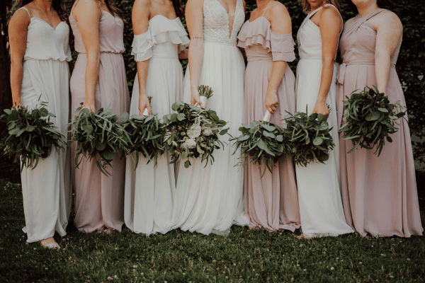 Bride and bridesmaids in white and blush dresses standing outdoors with greenery bouquets.