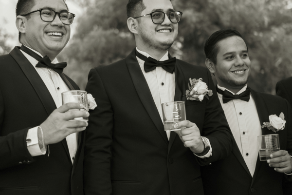 Three groomsmen wearing black tuxedos and bow ties, holding drinks and smiling outdoors.
