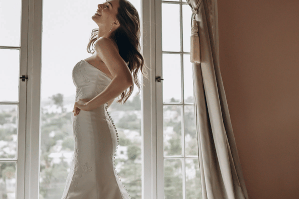 Bride wearing a fitted satin wedding dress with a corset back near a window.