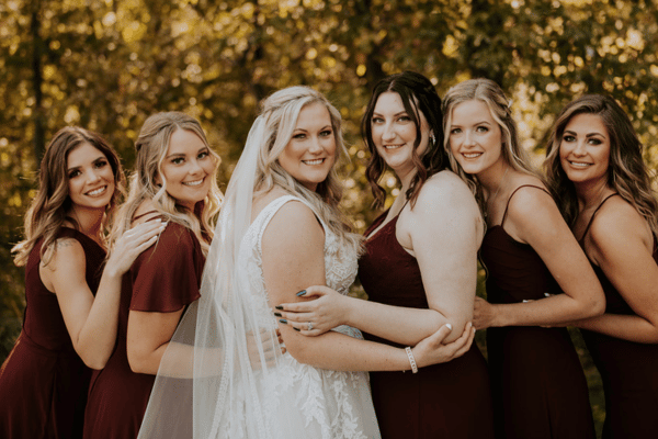 Bride in a lace wedding dress posing with her bridesmaids outdoors in a wooded setting.